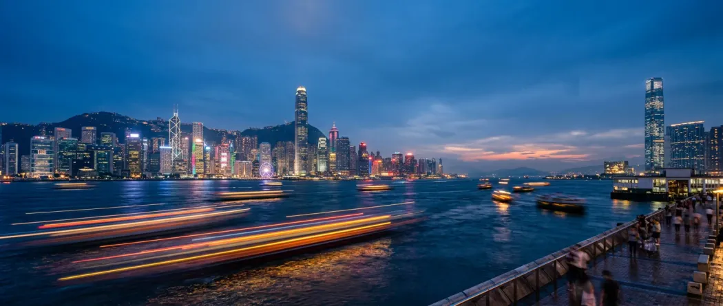 Hong Kong skyline at blue hour from across Victoria Harbour with neon orange light trails crossing the water toward mainland China